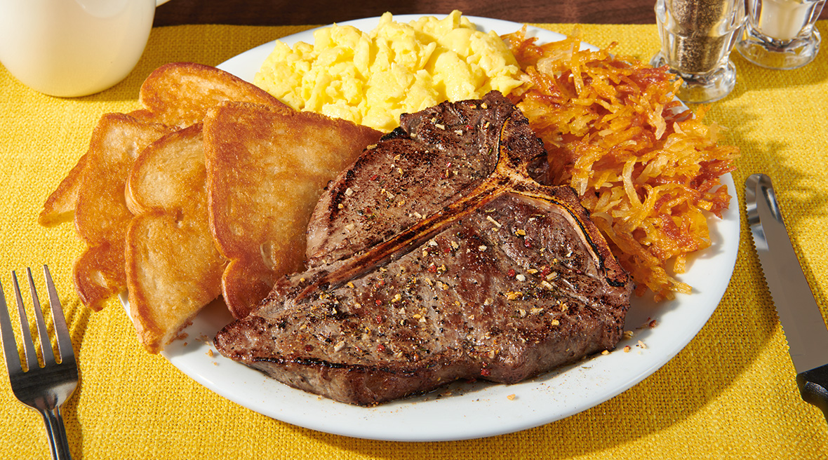 Hearty plate with a big T-bone steak served with fluffy scrambled eggs, toast, and crispy hash browns on the side.