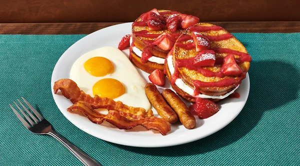 Fluffy brioche French toast stuffed with sweet cream and drizzled with berry sauce on a plate with eggs, bacon, and sausage links.