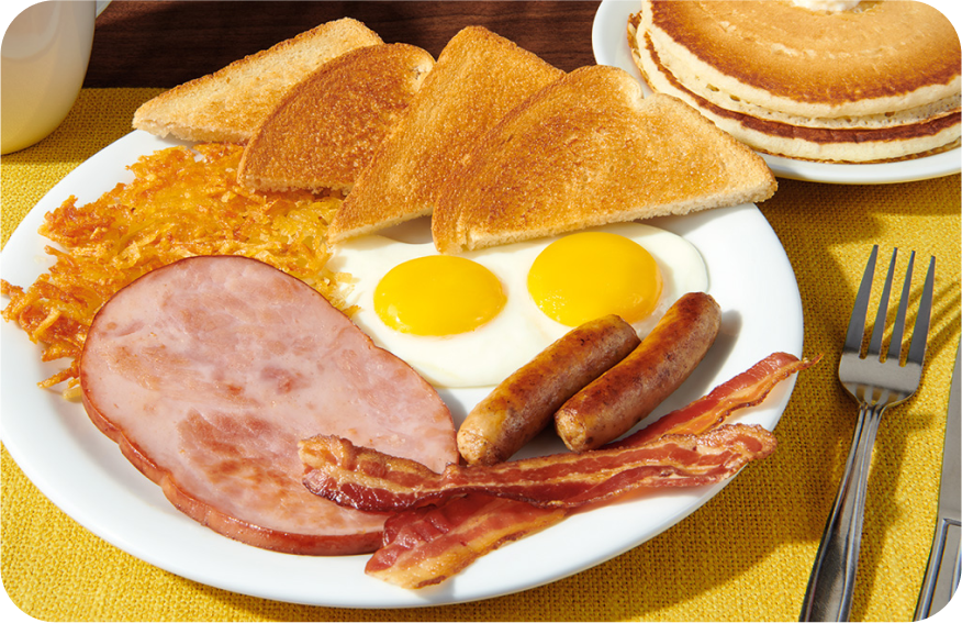 Grilled ham, sausage links, savory bacon, crispy hash browns, eggs and toast on a plate with a side stack of pancakes. 