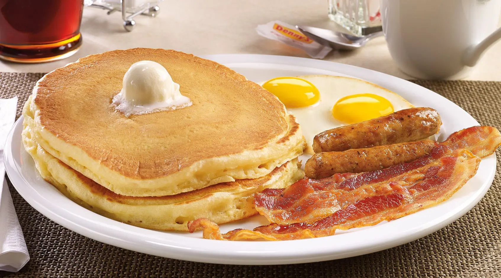 A classic slam breakfast plate with stacked pancakes topped with butter, sausage links, bacon, and a pair of sunny-side-up eggs.