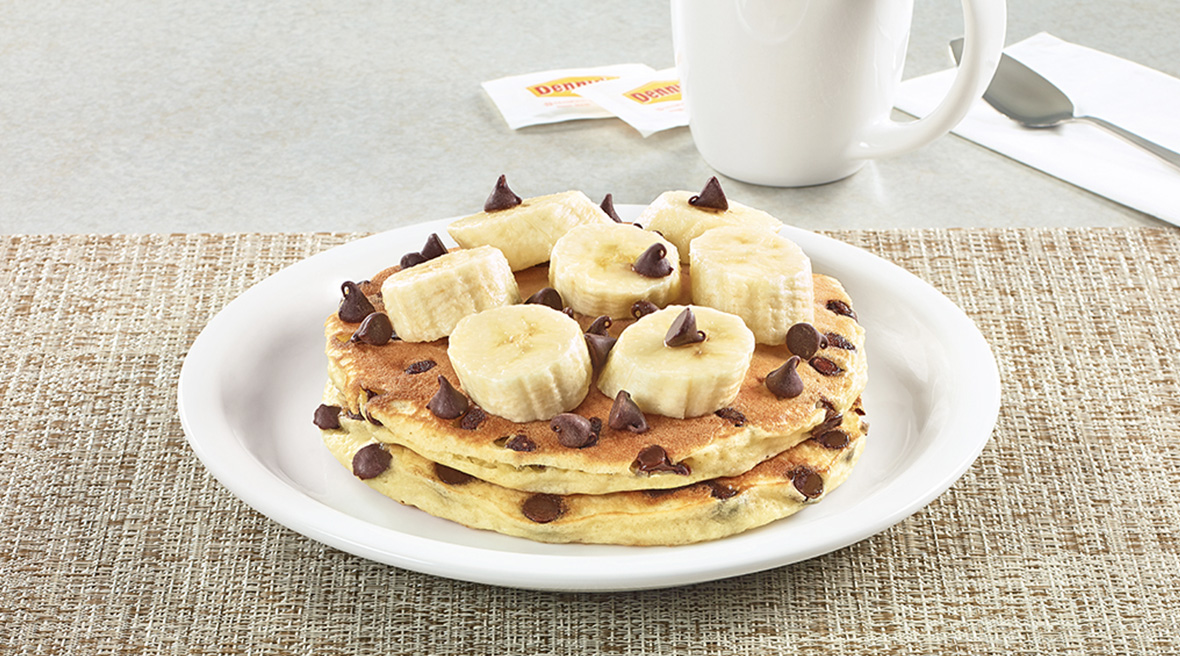 A double stack of chocolate chip pancakes generously topped with sliced bananas and sweet chocolate morsels.