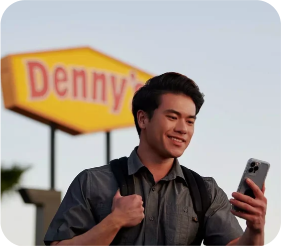 A man smiles and walks to a Denny's diner while looking at the Denny's App on his phone. 