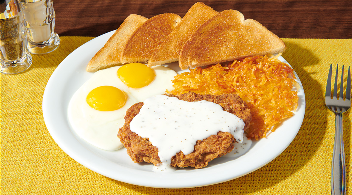 Breakfast plate with country fried steak smothered in white gravy along with a side of crispy hash browns, toast, and sunny-side-up eggs.